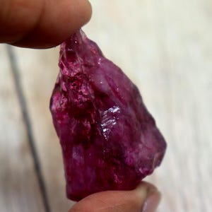 May include: A raw ruby gemstone, held between fingers. The uncut gemstone is a deep magenta hue with a rough, textured surface. The ruby has an irregular shape and reflects light, highlighting its crystalline structure. A natural, unpolished gemstone.