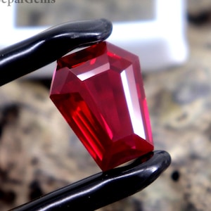 Puede incluir: Una gema roja intensa, tallada en forma rectangular, es sostenida por unas pinzas negras sobre un fondo texturizado de tonos neutros. La gema, posiblemente un rubí, tiene un alto brillo. La imagen está bien iluminada, resaltando las facetas.
