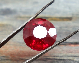May include: A deep red ruby gemstone with a faceted cut, held by tweezers against a woodgrain background.