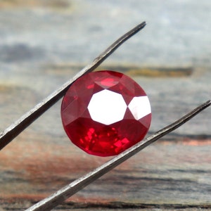 Puede incluir: Una piedra preciosa de rubí rojo intenso con un corte facetado, sostenida por unas pinzas sobre un fondo de madera.