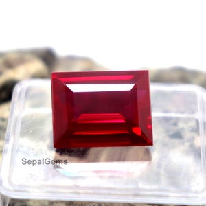Peut inclure: Une pierre précieuse rouge de taille rectangulaire aux surfaces facettées, posée sur un présentoir en plastique transparent. La pierre présente une teinte cramoisie profonde et saturée, avec le texte SepalGems inscrit sur la base.
