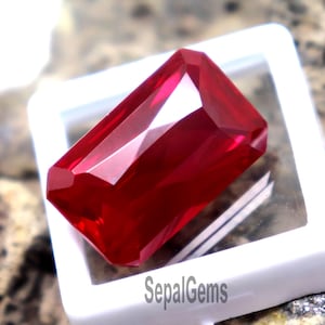 May include: A vibrant, rectangular, faceted red gemstone displayed in a square white plastic case with a clear lid. The case features the text SepalGems printed in black on the lower edge, showcasing the gemstone's deep crimson color and clarity.
