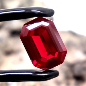 May include: A faceted, rectangular ruby gemstone held by black tweezers. The gem is a deep, vibrant red colour, with sharp, clean edges. The background is blurred, with the text "SepalGems" visible.