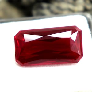 Puede incluir: Una gema de color rojo intenso y vibrante con un corte esmeralda rectangular, que presenta múltiples facetas que reflejan la luz. La gema está colocada sobre una base de espuma blanca dentro de una caja de plástico transparente y cuadrada.