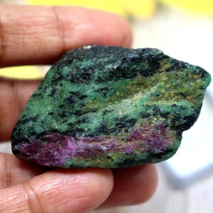 Könnte beinhalten: Ein roher Edelstein mit einer Mischung aus Farben. Der Stein hat eine grüne Basis mit schwarzen Sprenkeln und Flecken von Lila und Gelb. Der Stein wird in einer Hand gehalten.