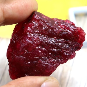 May include: A rough, uncut ruby gemstone with a deep red color. The ruby is held between fingers, showcasing its irregular shape and natural texture. The background is blurred, highlighting the gemstone's rich hue and raw beauty.