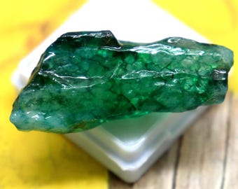 May include: A raw, elongated emerald gemstone with a deep green hue and a textured, crystalline surface. The stone features natural inclusions and facets, resting horizontally on a small, square, white plastic display base for presentation.