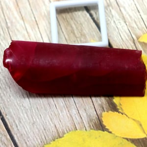 May include: A dark red, cylindrical object with a textured surface, possibly a candle or decorative item. A white square frame sits behind it, with yellow leaves on a wooden surface.