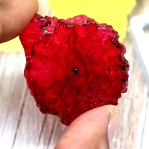 Könnte beinhalten: Eine leuchtend tiefrote, kreisförmige Scheibe aus getrockneter Drachenfrucht mit einer texturierten, kristallinen Oberfläche und einem kleinen, dunklen Loch in der Mitte. Die Fruchtscheibe hat einen gezackten Rand und wirkt rubinrot.