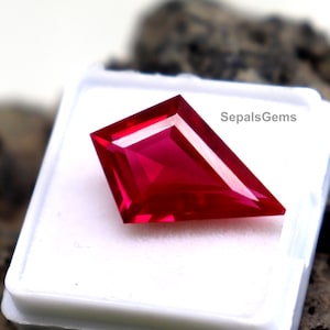 Puede incluir: Una piedra preciosa roja facetada en forma de cometa con un acabado brillante y transparente. La piedra se muestra dentro de un pequeño recipiente cuadrado de plástico blanco con un inserto de espuma blanca. El texto SepalsGems está impreso en negro.
