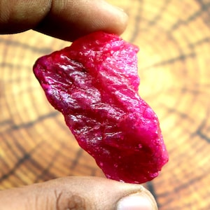 May include: A raw, uncut ruby gemstone with a deep, vibrant magenta-red color. The stone features a natural, irregular, and textured surface with translucent areas that catch the light, showcasing its crystalline structure and organic shape.