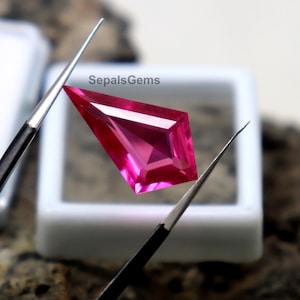 Puede incluir: Una piedra preciosa facetada en forma de cometa de color rosa intenso, sostenida por dos pinzas metálicas finas. La piedra presenta ángulos geométricos afilados y un corte brillante. El texto SepalsGems es visible en el fondo.
