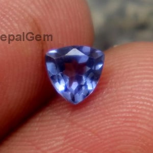 Könnte beinhalten: Ein dreieckiger, facettierter, blauer Edelstein. Der Edelstein wird zwischen Fingern gehalten, wodurch seine Form und Farbe zur Geltung kommen. Der Stein hat eine polierte Oberfläche.