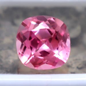 Puede incluir: Una gema rosa facetada, con corte cojín, exhibida en una caja de plástico blanco. La gema tiene un tono rosado vibrante y una superficie brillante y reflectante. La caja proporciona un fondo limpio y neutro, resaltando el color y el corte de la gema.