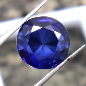 May include: A round, faceted, deep blue gemstone held by silver tweezers. The gem has a brilliant cut, reflecting light to create a starburst effect. The background is a blurred white square and a neutral surface.