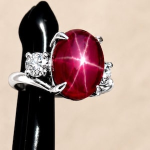 Puede incluir: Un anillo de plata con una gema grande, ovalada, de color rojo intenso con un patrón de estrella de seis puntas. El anillo está flanqueado por dos gemas más pequeñas, redondas y transparentes. El anillo se exhibe sobre un soporte negro.