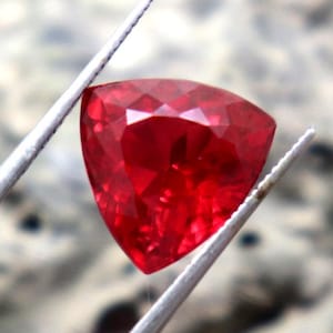 Könnte beinhalten: Ein leuchtend roter Edelstein mit einem dreieckigen Schliff, gehalten von einer Pinzette vor einem verschwommenen Hintergrund.