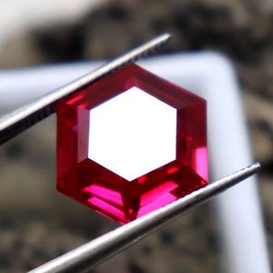 May include: A deep red hexagonal gemstone held by tweezers. The gemstone is faceted and has a high polish.