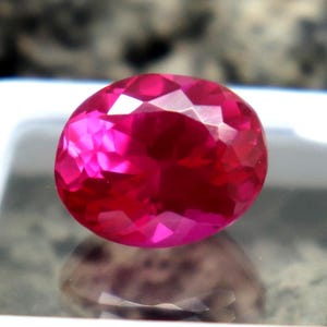May include: A faceted, oval-shaped, vibrant pink gemstone. The gem is displayed in a white square container, reflecting light and color. The background is a blurred, textured surface.