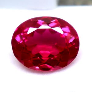 May include: A faceted, oval-shaped, ruby-red gemstone. The gem is set against a white background within a white plastic case. The gemstone's facets reflect light, creating a brilliant display of color and sparkle. This gemstone is suitable for jewelry making.