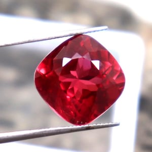 Könnte beinhalten: Ein tiefroter Edelstein mit einem Kissen-Schliff. Der Edelstein wird von einer Pinzette gehalten und ist scharf vor einem verschwommenen Hintergrund.