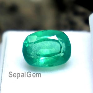 May include: A bright green, oval-shaped emerald gemstone presented in a white plastic case. The gem's facets are visible, reflecting light. The text "SepalGem" is printed on the case. The background is out of focus.