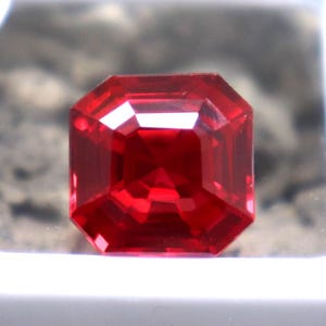 Puede incluir: Una gema roja vibrante con un corte cuadrado esmeralda, exhibida en una caja cuadrada blanca. La gema es de un color rojo intenso y rico y tiene un alto nivel de claridad.