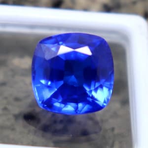 Puede incluir: Una vibrante piedra preciosa de zafiro con talla cojín, exhibida en una caja de plástico blanco. El zafiro exhibe un tono azul profundo y rico con múltiples facetas que reflejan la luz. La piedra preciosa se encuentra sobre un fondo neutro.