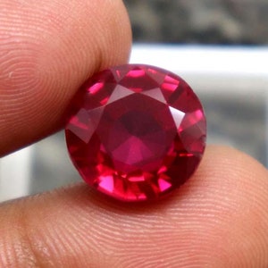 May include: A deep red gemstone with a round, faceted cut. The gemstone is held in a hand, showcasing its size and brilliance.