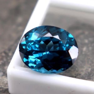 Puede incluir: Una gema facetada, de forma ovalada y color verde azulado se muestra en una caja de pl&aacute;stico blanca. La gema tiene un corte brillante, que refleja la luz. El fondo es una superficie gris texturizada. Esta gema es para joyer&iacute;a.