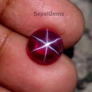 May include: A polished, deep red gemstone with a six-rayed star pattern. The gem is held in a hand, highlighting its vibrant colour and unique optical effect. The text "SepalGems" is visible.