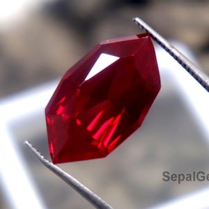 May include: A faceted, deep red gemstone is held by tweezers. The gem has a vibrant, saturated colour and multiple reflective surfaces. The image includes the text "SepalGems".