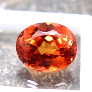 Pode incluir: Uma gema facetada de forma oval com tons vibrantes de laranja e amarelo. A gema é apresentada numa caixa de exposição branca, realçando o seu brilho. As facetas da gema refletem a luz, criando um efeito cintilante. Ideal para joalharia.