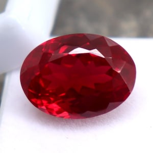 Peut inclure: Une pierre précieuse rouge foncé, de forme ovale et à facettes. La gemme est posée sur une surface blanche, avec une structure blanche visible en arrière-plan. Les facettes de la pierre reflètent la lumière.