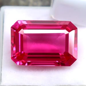 May include: A vibrant, rectangular-cut, hot pink gemstone. The gem is set in a white display case, with facets catching the light. The gemstone's color is a deep, saturated pink, with a polished, reflective surface. The gem is likely a ruby or pink sapphire.