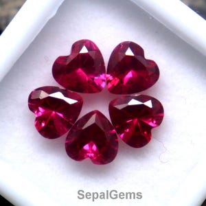 May include: Five heart-shaped, faceted, deep red gemstones arranged in a flower-like pattern inside a white square display case. The gemstones are set against a blurred, textured background. The text "SepalGems" is visible.