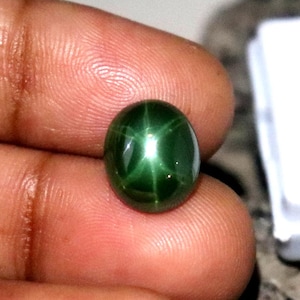 Pode incluir: Uma pedra preciosa ovalada verde-escura polida com um padrão em forma de estrela. A gema é segurada entre os dedos, com uma caixa branca e o texto "SepalGems" no fundo. O padrão de estrela é uma característica marcante.