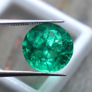 Pode incluir: Uma gema de esmeralda verde vibrante mantida em pinças. A esmeralda é redonda e tem um corte facetado.