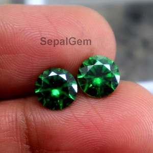 May include: Two round, faceted, emerald green gemstones. The gems are held in a hand, with a white box in the background. The gems have a brilliant cut, reflecting light.
