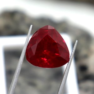 May include: A deep red triangular gemstone held by tweezers. The gemstone is faceted and has a high shine.
