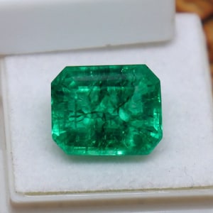 May include: A vibrant green emerald gemstone with a rectangular cut, showcasing intricate internal inclusions. The emerald is displayed in a white velvet-lined jewelry box.