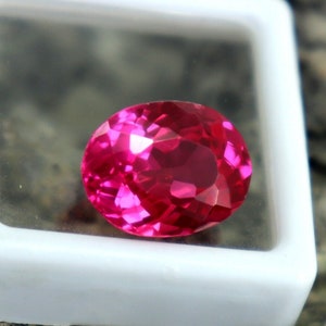 May include: A vibrant pink oval-shaped gemstone with a faceted cut. The gemstone is displayed in a clear plastic box.