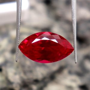 Könnte beinhalten: Ein facettierter Rubin im Marquise-Schliff, gehalten von einer Pinzette. Der Rubin hat eine leuchtend rote Farbe, mit vielen Facetten, die das Licht reflektieren. Die Form des Edelsteins ist länglich und an beiden Enden spitz.