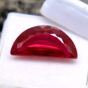 May include: A deep red, half-moon shaped gemstone with a faceted cut, displayed in a white jewellery box.