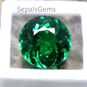 May include: A round, faceted, emerald-green gemstone is displayed in a white square case. The gemstone has a brilliant cut, reflecting light. The text "SepalsGems" is visible on the case. The background is a textured, neutral-toned surface.