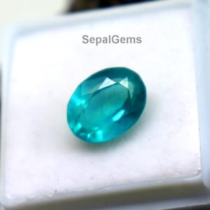 Puede incluir: Una gema ovalada de color verde azulado se muestra en una caja de plástico blanco con tapa transparente. La gema está sobre un fondo blanco. La caja descansa sobre una superficie rugosa de color marrón oscuro. El texto "SepalGems" es visible.
