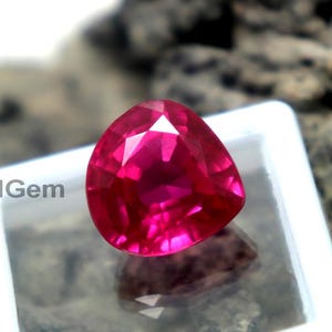 Può includere: Una gemma rosa vibrante, sfaccettata e di forma triangolare, è esposta in un contenitore quadrato bianco. La gemma è posta su uno sfondo strutturato grigio e marrone. Le sfaccettature della gemma riflettono la luce, creando un effetto scintillante.