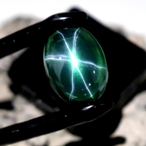 Pode incluir: Uma pedra preciosa verde oval polida com um padrão em forma de estrela, segurada por pinças pretas. A gema está sobre um fundo escuro e repousa sobre uma rocha áspera e texturizada. O texto "SepalGems" é visível.