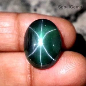Pode incluir: Gema ovalada verde escuro com um padrão de estrela de seis pontas. A gema tem uma superfície polida e um efeito de estrela proeminente, com linhas brancas irradiando do centro. A gema é segurada em uma mão.