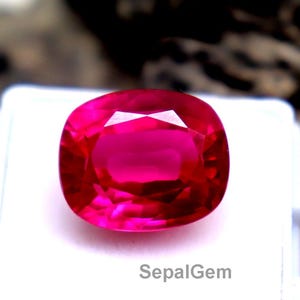 Puede incluir: Un vibrante rubí tallado en cojín, presentado sobre una superficie blanca. El rubí es de un rojo intenso y saturado con facetas que capturan la luz. El texto "SepalGem" está impreso en la superficie blanca. El fondo es una superficie texturizada borrosa.